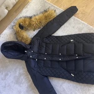 Svart rockanblue jacka  - Svart lång pufferjacka med quiltade partier och avtagbar huva. Jackan har dragkedjor på fickorna och en snygg fuskpälsdetalj som kan fästas på huvan. Perfekt för kalla dagar och har en stilren look.