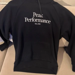 Svart hoodie från Peak Performance - Svart hoodie från Peak Performance med vit logotyp på bröstet och klassisk magficka. Tröjan har huva med snörning och långa ärmar, perfekt för chill dagar eller när du vill ha en clean streetstyle-look.