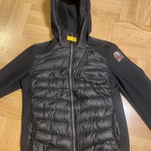 Svart cardigan jacka från Parajumpers - Svart pufferjacka från Parajumpers med huva, quiltad framsida och dragkedja. Jackan har en gul detalj vid halsen och en patch på ärmen. Perfekt för kyliga dagar och har en sportig vibe.