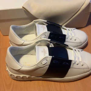 Valentino Garavani vita sneakers - Snygga vita sneakers från Valentino Garavani med bred svart rem över vristen och coola nitar på hälen. Skorna har rund tå, platt sula och är tillverkade i skinn. Perfekta för dig som vill ha en stilren men edgy look.