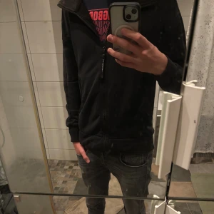 Svart zip up tröja från C.P. Company - Svart zip up från C.P. Company med dragkedja framtill och två fickor. Jackan har huva med de klassiska C.P. Company-glasögonen. 