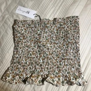 Blommig tubtopp från Design by Si - Supersöt tubtopp med smock och volangkant från Design by Si. Toppen har ett blommigt mönster i beige, grönt och brunt på vit botten. Aldrig använd. Storleken står inte men tror att det är storlek XS/S💋