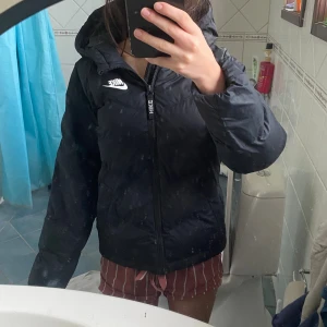 Svart Nike jacka  - Svart pufferjacka från Nike med vit logga på bröstet och huva. Jackan har quiltad design, dragkedja framtill och långa ärmar. Perfekt för kalla dagar och har en clean, sportig look som funkar till streetwear. Materialet är syntet och jackan är lätt vadderad.