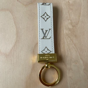  Vuitton vit nyckelring i skinn - Exklusiv nyckelring från  Vuitton i vitt skinn med klassiskt monogrammönster i guld. Detaljer i guldfärgad metall och präglad logotyp på ringen. Perfekt accessoar för att lyfta din väska eller nycklar med lyxig känsla.