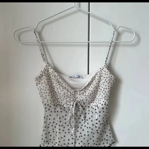 Prickig linnetopp från Bershka - Supersöt linnetopp från Bershka i vit med bruna prickar. Tunn och luftig med smala axelband och snygg snörning framtill som ger en cool detalj. Perfekt för varma dagar och enkel att styla med jeans eller kjol.
