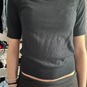 Svart stickad topp med kort ärm - Snygg svart stickad topp med korta ärmar och rund hals. Modellen är croppad och har ribbad kant nedtill för en schysst passform. Perfekt att matcha med höga jeans eller kjol för en clean look.