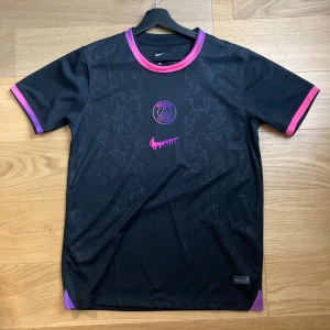 PSG Nike Edtion T-shirt - Svart PSG fotbollströja från Nike med lila och rosa detaljer. Tröjan har korta ärmar, diskret blommönster i tyget och en unik rosa Nike-logga med drip-effekt. Tillverkad i Dri-FIT-material som andas, perfekt för träning eller match.