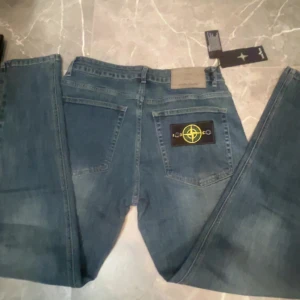 Stone Island blå jeans med patch - Snygga blå jeans från Stone Island med klassisk gul och grön kompass-patch på bakfickan. Jeansen har raka ben, normal passform och är tillverkade i slitstarkt denim. Fina detaljer med märkets logga på både etikett och patch. Perfekt för dig som gillar streetwear.