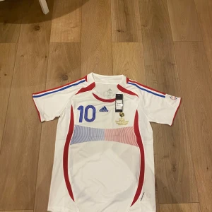 Frankrike Zidane #10 fotbollströja Adidas - Säljer en officiell fransk landslagströja från Adidas med Zidane och nummer 10 på ryggen. Tröjan är vit med röda och blå detaljer, korta ärmar och tryckta ränder på axlarna. Materialet är lätt polyester, perfekt för match eller träning.