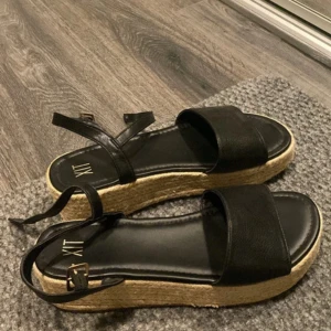 Svarta platåsandaler från XIT - Snygga svarta sandaler från XIT med bred rem över tårna och justerbart spänne vid ankeln. Skorna har en rejäl platåsula i flätad jute-look som ger extra höjd och sommarkänsla. Perfekta till jeans eller klänning för en trendig vibe.