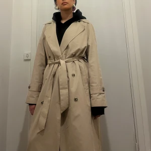 Beige trenchcoat med bälte - Snygg beige trenchcoat med klassisk dubbelknäppning och bälte i midjan. Jackan har långa ärmar, breda slag och knappar i fram. Använd 2 gånger NY PRIS: 1199. Mitt pris är inte hugget i sten