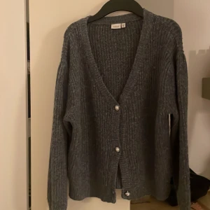 Mörkgrå ribbad kofta från Name It - Mysig mörkgrå kofta från Name It i ribbstickad design. Koftan har v-ringad hals, tre pärlemorknappar framtill och långa ärmar. Perfekt för att slänga över en t-shirt när det är lite kyligt ute.