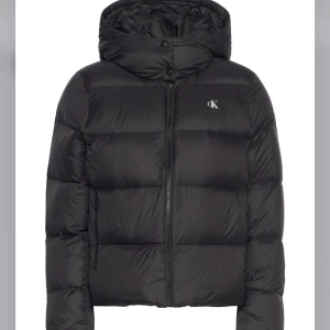 Svart pufferjacka från Calvin Klein - Snygg svart pufferjacka från Calvin Klein med huva och diskret CK-logga på bröstet. Jackan har dragkedja framtill och är vadderad för extra värme. Perfekt för kalla dagar och har en modern, boxig passform.