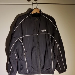 Svart vindjacka från FSBN Chaotic  - Svart vindjacka från Chaotic med vita kontrastlinjer. Elastiska muddar och två fickor med dragkedja. Sportig look med logga på bröstet och  detaljer längs ärmarna.