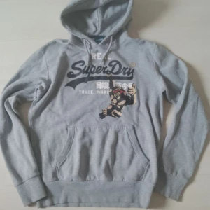Grå Superdry hoodie med tryck - Snygg grå hoodie från Superdry med stort tryck på bröstet och cool gorilla med röda solglasögon. Tröjan har huva med snörning och en stor magficka. Perfekt för dig som gillar streetstyle och vill sticka ut lite extra.