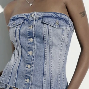 Blå denim korsett-tubtopp med knappar - Säljer en snygg blå denim korsett-tubtopp från Zara. Toppen är axelbandslös med knappar framtill, två dekorativa fickor med knappdetaljer och rå kant nedtill. Den har en tight passform och cool jeanslook som ger en edgy vibe.