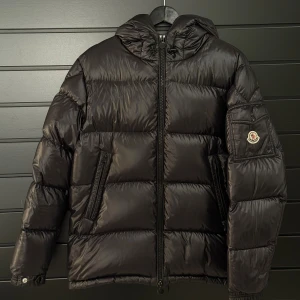 Svart Moncler pufferjacka med huva - Svart pufferjacka från Moncler som jag säljer åt min kille då han inte riktigt får användning av den. Är i modellen ”Ecrins Giubbotto” i storlek 1 (motsvarar en S). Är lite sliten vid ett visst ställe som även visas på bilden. 100% äkta såklart och går att få fler bilder om de behövs ☺️