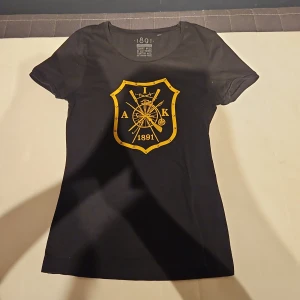 Svart AIK limited edition t-shirt - Svart t-shirt från 1891 med AIK:s klassiska klubbmärke i gult på bröstet. Limited edition, nummer 54 av 200. T-shirten har rund hals, korta ärmar och är gjord i mjuk bomull. Perfekt för dig som vill visa ditt AIK-stöd med stil.