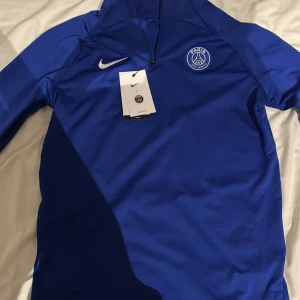 Blå Nike PSG half zip träningströja - Snygg blå träningströja från Nike och PSG med half zip och broderad klubbmärke på bröstet. Slim fit-modell i Dri-Fit polyester som andas, perfekt för fotboll eller sport. Långärmad med vit Nike-logga och coolt Paris Saint-Germain-emblem.