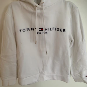 Vit hoodie från Tommy Hilfiger  - Vit hoodie från Tommy Hilfiger med broderad logga och text på bröstet. Klassisk huva med snörning och långa ärmar. Mjuk och skön bomullsblandning, perfekt för chill dagar. Liten flaggdetalj på ärmslutet för extra stilpoäng.