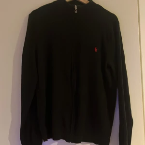 Svart kofta från Polo Ralph Lauren -  Tja säljer en Svart kofta från Polo Ralph Lauren med dragkedja framtill och klassisk röd broderad logga på bröstet. Tröjan är långärmad och har en stilren, enkel design!