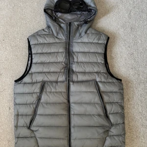 Grå dunväst med huva från C.P. Company - Snygg grå dunväst från C.P. Company med ikoniska goggles i huvan. Västen har quiltad design, dragkedja framtill och två stora fickor. Perfekt för lager-på-lager och streetwear-vibe. Materialet är lätt och håller värmen utan att bli klumpig. Lite stor i storlek 