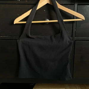 Svart croppad halterneck linnetopp med breda axelband - Säljer en svart croppad linnetopp med breda axelband och fyrkantig halsringning. Toppen har en enkel och stilren design med lätt rynkning framtill. Perfekt för dig som gillar minimalistisk stil och vill ha en topp som är lätt att matcha.