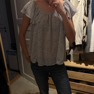 Vit blommig blus med volangärm - Söt och luftig blus i vitt med små svarta prickar. Blusen har korta volangärmar och en lös passform med rundad halsringning. Perfekt för varma dagar och enkel att styla med jeans eller kjol.