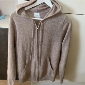 Beige hoodie i kashmir från John Henric - Mysig beige hoodie i lyxig kashmir från John Henric. Tröjan har dragkedja framtill, huva med snörning och två fickor. Perfekt för dig som gillar en stilren och avslappnad look med premiumkänsla.