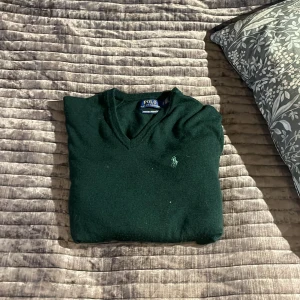 Mörkgrön merinoullströja Polo Ralph Lauren - Snygg mörkgrön stickad tröja från Polo Ralph Lauren i mjuk merinoull. V-ringad modell med klassisk broderad logga på bröstet. Perfekt för dig som gillar stilrena och tidlösa plagg med skön passform.