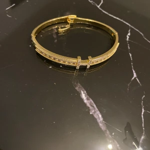 Guldigt armband med kristaller - Snyggt guldigt armband med en rad glittrande kristaller och dekorativt spänne täckt av små stenar. Armbandet har en stilren och modern design som verkligen sticker ut. Perfekt för dig som gillar bling och vill ha något som syns.