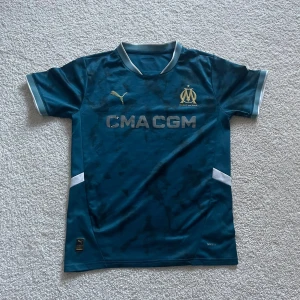 Blå Marseille fotbollströja Puma - Säljer en snygg blå Olympique de Marseille fotbollströja från Puma med korta ärmar och silverfärgade detaljer. Tröjan har klubbens emblem och sponsortryck på bröstet, samt ljusblå och vita inslag vid krage och ärmslut. Tillverkad i lätt och ventilerande polyester.