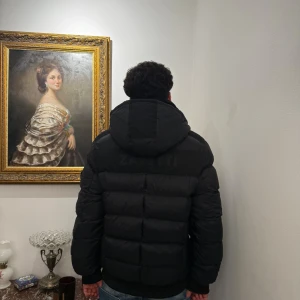 Svart pufferjacka från Zavetti Canada - Säljer en svart pufferjacka från Zavetti Canada med huva och dragkedja framtill. Jackan har en broderad lönnlövpatch på ärmen och flera praktiska fickor. Materialet är polyamid och polyester, perfekt för kalla dagar. Snygg och stilren modell som håller värmen.