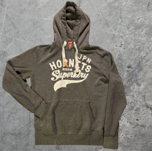 Superdry hoodie - Säljer en grå hoodie från Superdry med stort vitt tryck på bröstet och japanska tecken. Tröjan har huva med snörning, känguruficka och mjukt foder. Perfekt för dig som gillar streetwear och vill ha en skön och snygg tröja till vardags. Om du undrar något är det bara att skriva, priset är inte hugget i sten✅🙌