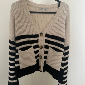 Randig kofta från beige och svart - Superfin ribbstickad kofta i beige och svart med breda ränder. Koftan har v-ringning, stora knappar framtill och två rejäla fickor. Långärmad med slits på ärmarna.