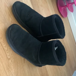 Svarta boots i mocka med rund tå - Svarta boots med rund tå och platt sula, tillverkade i mjuk mocka med värmande foder. Klassisk enkel design utan synliga märken, perfekt för kalla dagar. Skorna har en låg skaft och är helsvarta med diskreta sömmar.