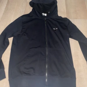 Svart hoodie från Comme des Garçons - Svart hoodie med dragkedja från Comme des Garçons Play. Har ett stort hjärta med  tryckt på ryggen och ett mindre hjärta på bröstet. Utrustad med luva och fickor framtill. Hör av dig för flera frågor. 