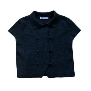 Svart kortärmad blus - ZARA - Kortärmad svart stickad tröja från Zara med klassisk krage och dubbla rader klädda knappar framtill. Toppens material är mjukt och har en lätt struktur, perfekt för en stilren look. Modellen är rak och något croppad.