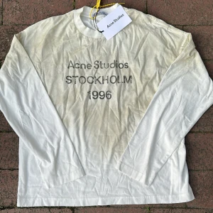Vit långärmad Acne Studios tröja - Vit långärmad tröja från Acne Studios med tryck 'Acne Studios STOCKHOLM 1996' på bröstet. Tröjan har rund hals och är gjord i mjuk bomull. Perfekt för dig som gillar clean och stilren design med en touch av street.                                                    Är inte helt säker om den är äkta för har köpt i andra hand  men den är väldigt snygg och i nytt skick!
