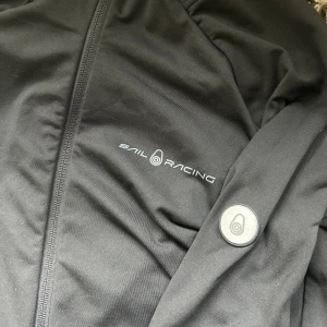 Svart jacka från Sail Racing - Svart jacka från Sail Racing med dragkedja framtill och logotyp på bröstet samt en rund logodetalj på ärmen. Jackan har en clean och stilren look,  perfekt för dig som gillar stilrena plagg. Materialet känns smidigt och lätt, med lång ärm och normal passform. Otroligt snygg badge som höjer upp hela outfiten. En jacka som passar de flesta stilarna såsom grisch. Storlek S. Nypris 2299 mitt pris 949. Priset går att diskutera vid snabb affär// Melvin
