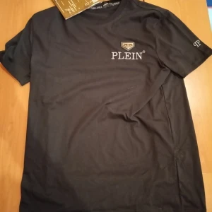 Svart Philipp Plein t-shirt med logga - Svart t-shirt från Philipp Plein med korta ärmar och snyggt broderad logga på bröstet samt en guldig detalj. Tillverkad i mjuk bomull och har PP-märke på ärmen. Perfekt för dig som gillar exklusiva streetwear-plagg. QR koderna för dig till Phillipp Pleins hemsida och den köptes för 250€. Tveka inte att höra av dig om du har några frågor eller liknande. Pris kan diskuteras.