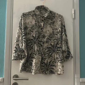 Beige skjorta med svart blommigt mönster - Snygg beige skjorta med svart blommigt och tropiskt mönster. Skjortan har klassisk krage, knappar framtill och långärmade ärmar. Perfekt för dig som gillar unika prints och vill sticka ut lite extra. Materialet känns mjukt och luftigt mot huden. Vet ej storlek gissar på L