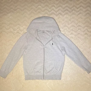 Ljusgrå hoodie från Polo Ralph Lauren - Snygg ljusgrå hoodie från Polo Ralph Lauren med dragkedja och klassisk logga broderad på bröstet. Tröjan har huva, två fickor fram och långa ärmar med ribbade muddar. Perfekt för chill dagar och enkel att matcha med jeans eller joggers. Pris kan diskuteras.