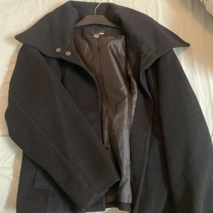 Svart rock från H&M med bred krage - Svart rock från H&M med bred krage och snygga knappar framtill. Jackan har två fickor på sidorna och är helfodrad med mjukt tyg på insidan. Klassisk och stilren modell som passar perfekt till kyliga dagar.