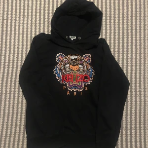 Svart Kenzo hoodie med tigerbrodyr XL - Svart hoodie från Kenzo i storlek XL med ikonisk broderad tiger och färgglada detaljer framtill. Tröjan har huva och långa ärmar, perfekt för dig som vill sticka ut med en streetstyle-look. Materialet är mjukt och skönt, troligen bomullsmix.