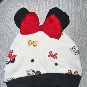 Mimmi Pigg mössa med öron och rosett - Supersöt mössa i vitt med svarta kanter, svarta öron och en stor röd rosett på toppen. Mössan har tryck med Mimmi Pigg, prickar och rosetter i rött, gult och svart. Perfekt för dig som älskar Disney och vill sticka ut lite extra. Från cirka 3 månader. Söt vit babymössa med svart kant och tryck av Musse Pigg-rosetter i olika färger. Mössan har även svarta öron och en stor röd rosett.