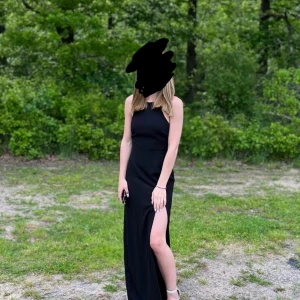 Svart långklänning med slits - Säljer en jättefin svart långklänning med hög slits på ena sidan. Klänningen är ärmlös och har en stilren, figurnära passform som ger en snygg siluett. Perfekt för dig som vill ha en klassisk look med lite edge. Den knyts i ryggen och den är använd 1 gång, hade den på balen i skolan