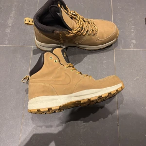 Nike Manoa boots i beige skinn - Nike Manoa boots i beige skinn med robust gummisula och snörning. Skorna har högt skaft, mörkbrunt foder och detaljer i guld på öglorna. Perfekta för höst och vinter med slitstark mocka och grovt mönstrad sula för extra grepp. Nypris 1249kr