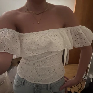 Vit offshoulder blus med volang - Superfin vit offshoulder blus med bred volang runt axlarna och broderade hålmönster. Sitter tight över midjan. Perfekt till ljusa jeans och smycken. Materialet känns som bomull och toppen är ärmlös med axlarna bara. Jag skickar snabbt