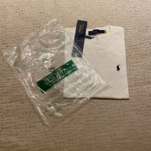Vit t-shirt från Polo Ralph Lauren - Vit klassisk t-shirt från Polo Ralph Lauren med rund hals och kort ärm. T-shirten har den ikoniska svarta polospelaren broderad på bröstet och är tillverkad i mjuk bomull. Perfekt basic-plagg med snygg passform och diskret logga.